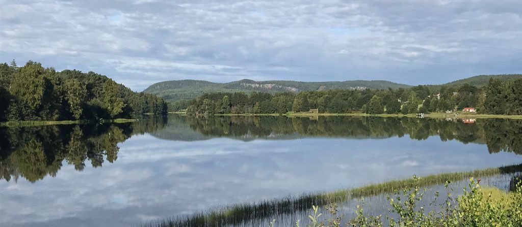 Bilden visar Dalälven en sensommardag sedd norrut från älvstranden i Gimsbärke. Den omgivande terrängen avspeglar sig i den lugna vattenytan. 