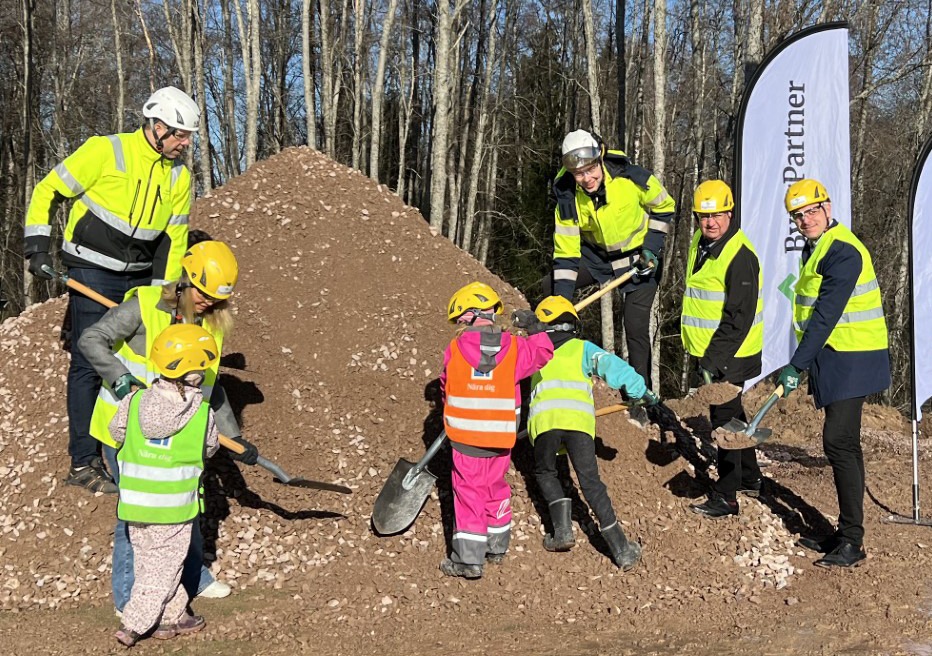 Fem vuxna personer och fem barn med spadar framför en grushög