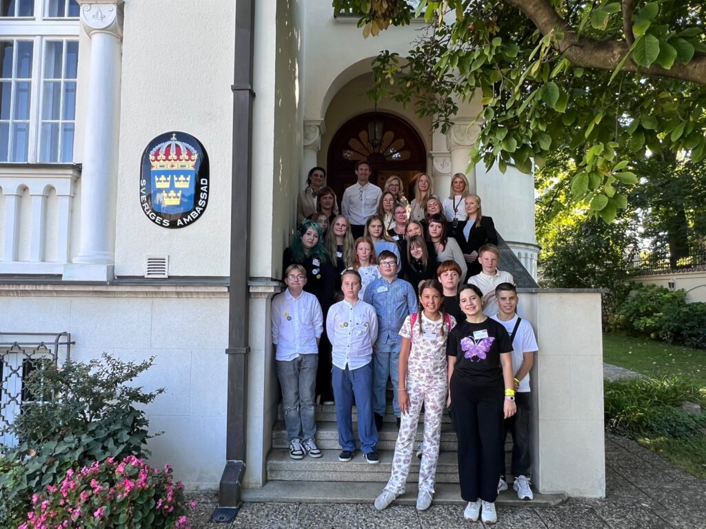 Ungdomar och vuxna på en trappa utanför en byggnad med en skylt för Sveriges ambassad.